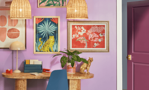 A room with walls painted light purple with eclectic framed prints and pendant lights along with a small desk.