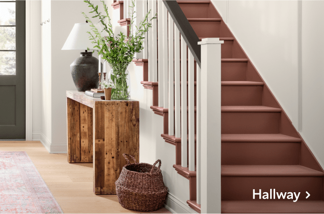 A hallway with white walls and natural accents next to a red-brown painted staircase.