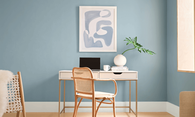 A tan wicker chair sits in front of a console table with a white and blue abstract print hung on a light blue-gray wall.