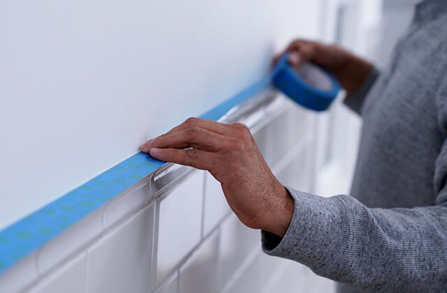 Person putting blue painters tape onto a white wall above tile.