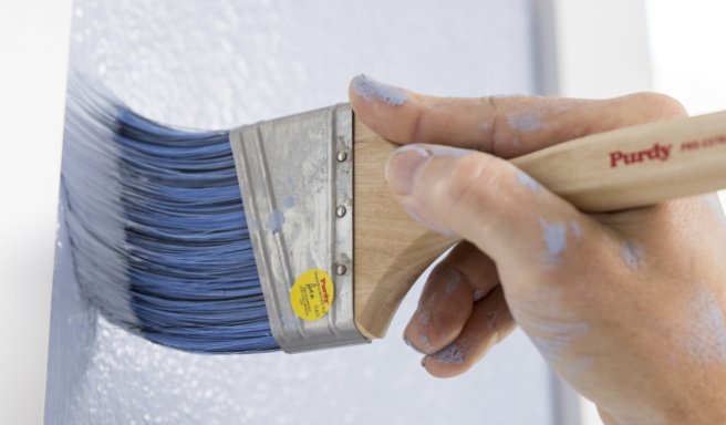 A person applying blue paint to a wall with a Purdy brush.