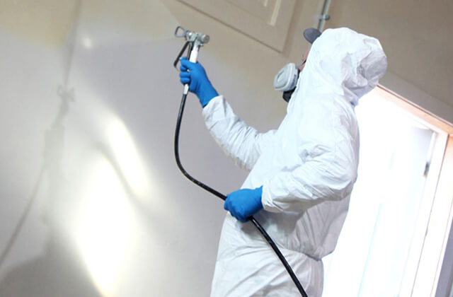 A person in a white coverall with blue gloves and respirator paint spraying a wall.