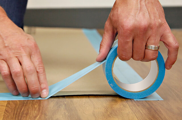 A person using blue painter's tape to tape down brown masking tape to a wood floor.