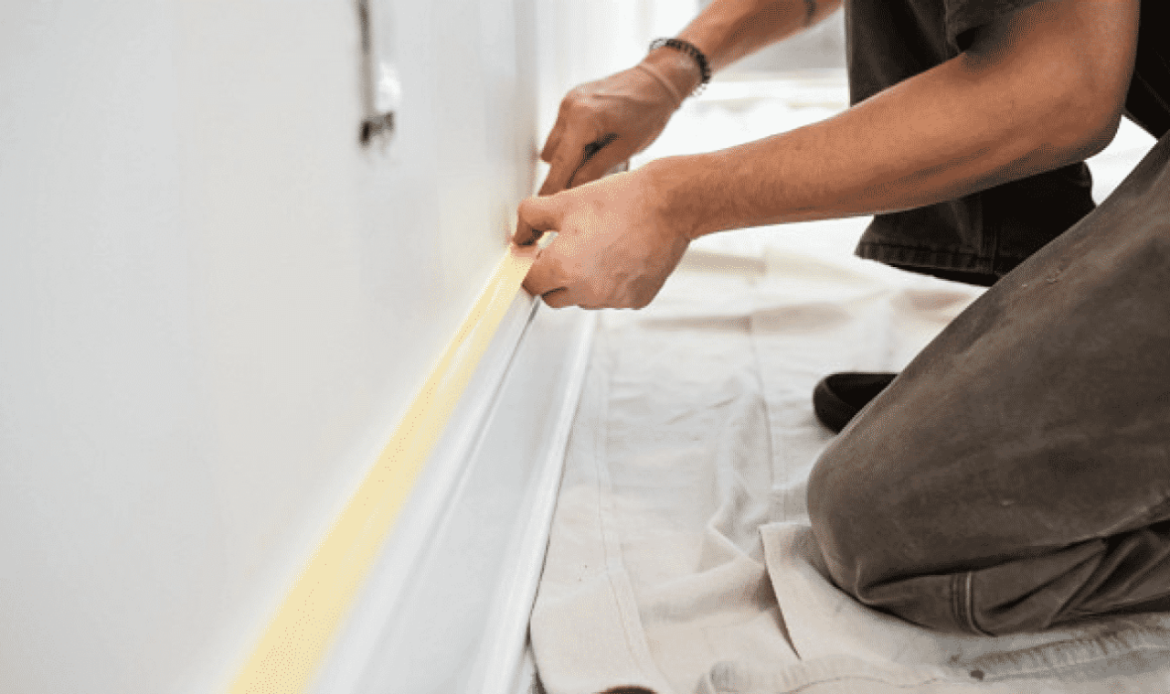 A person kneeling on a drop cloth applying masking tape to a white baseboard.
