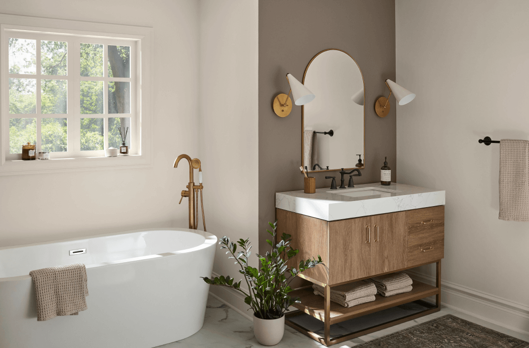 A bathroom with a window above a large white freestanding tub next to a plant and wooden vanity with marble counter under a mirror.