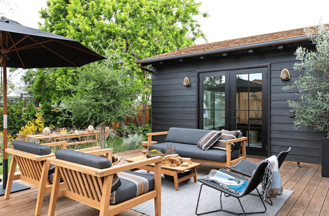 An outdoor deck space with two wooden chairs with cushions, wooden coffee table, wooden bench with cushions, two reclined chairs and an umbrella.