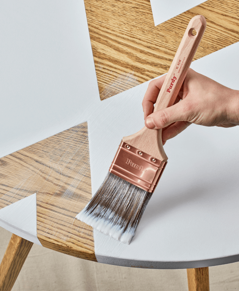 A person painting a wooden table with a Purdy paint brush.
