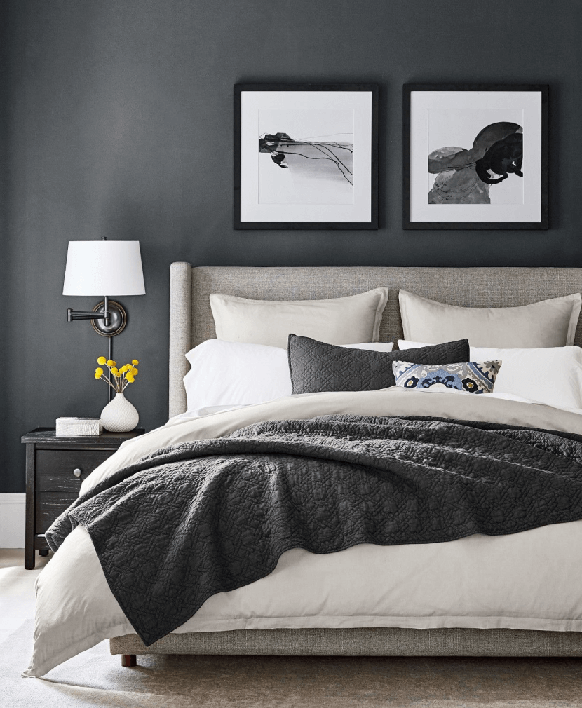 Bedroom with dark walls, grey headboard, grey and white pillows, bedside table with lamp and decor.