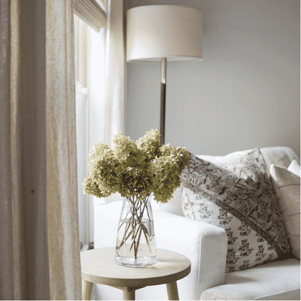 A vase of flowers on a table next to a white chair and lamp.