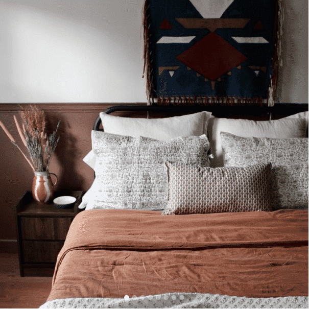 A bed with a tan comforter and 5 pillows next to a dark stained table, by theforestfarmhouse.