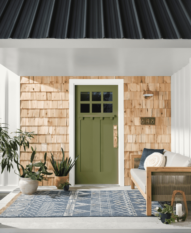 Front door painted green with wooden siding surrounding, couch and plants to fill the porch.