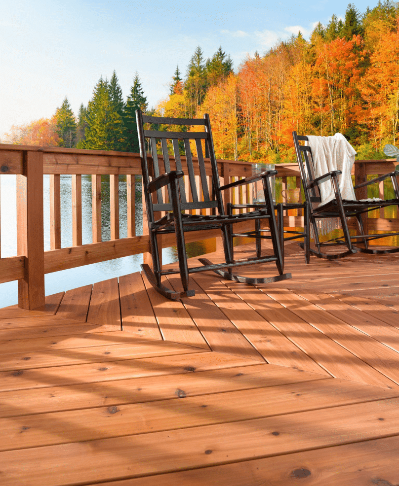 Wooden deck with black rocking chairs surrounding water and fall trees.