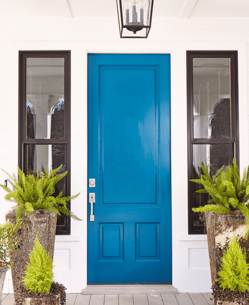 Bright blue front door in between two windows, surrounded by plants.
