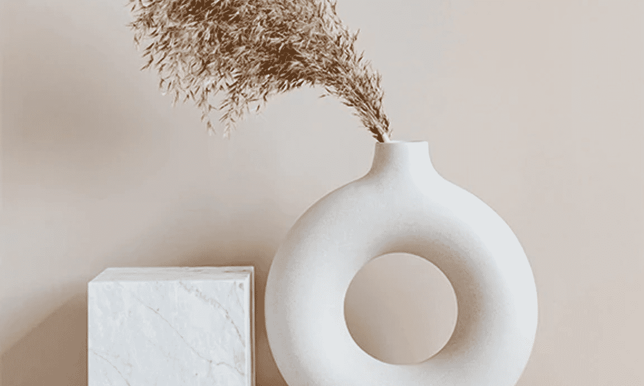 A cream circular vase with an open center with dried wheat next to a cube of granite.