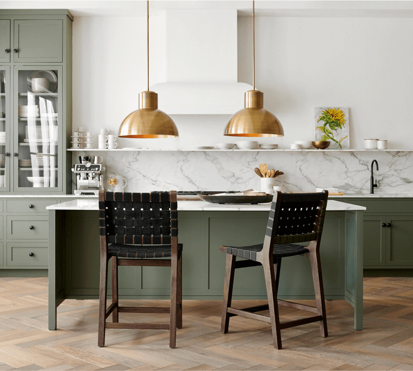 Bright modern kitchen with green painted cabinets, white walls, marble backsplash, gold lighting, and black barstools over wooden floors.