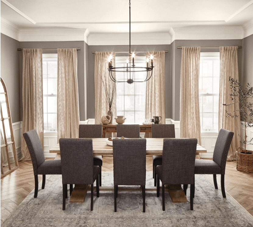 Neutral dining room with large windows, curtains, a chandelier above a large wooden table with cushioned chairs over an area rug.