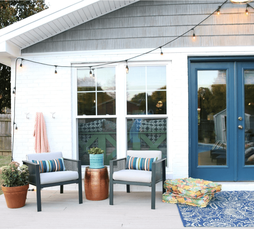 Outside of a house with blue glass doors, chairs, string lights, plants, and rug.