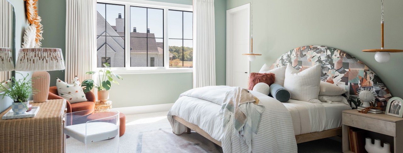 Soft green bedroom on a sunny day with lots of throw pillows and blankets on bed with oversized arch fabric-covered headboard, neutral accents and white curtains.