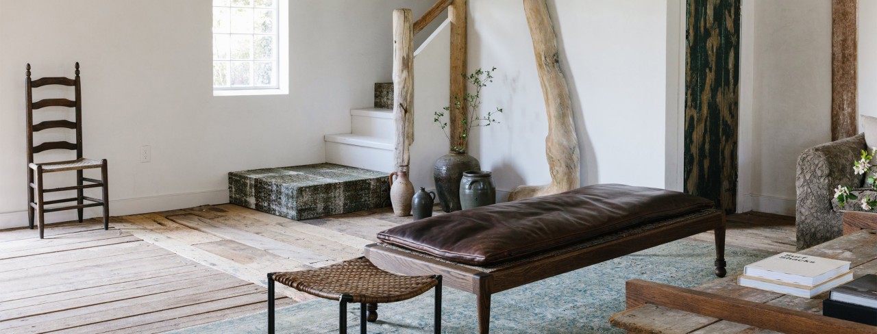 Interior of residential living space with white walls, natural wood beams and banisters in corner stairwell with collection of decorative ceramic vessels on the floor and leather-topped wood bench in foreground.