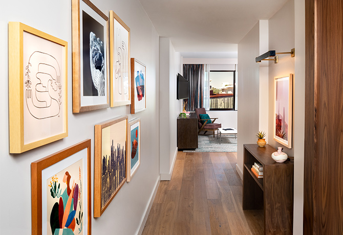 View down short hallway in hotel guest room, colorful gallery art on left wall, white walls and natural wood floors.
