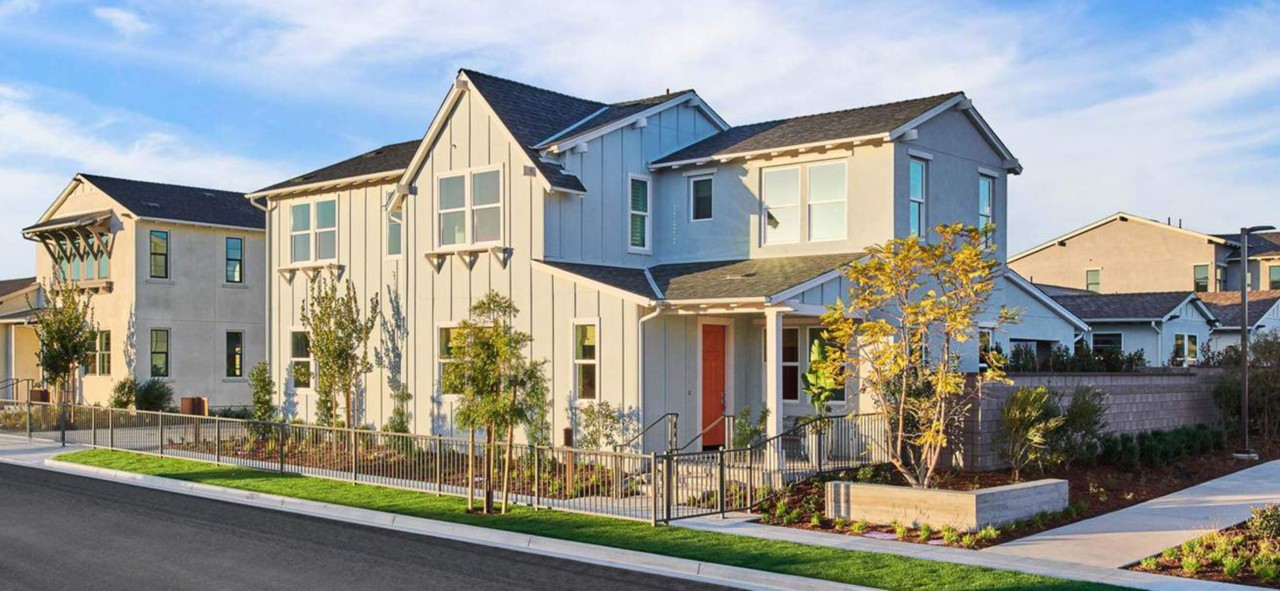 Angle shot of gray home exterior with dark orange front door and fence bordering sidewalk.