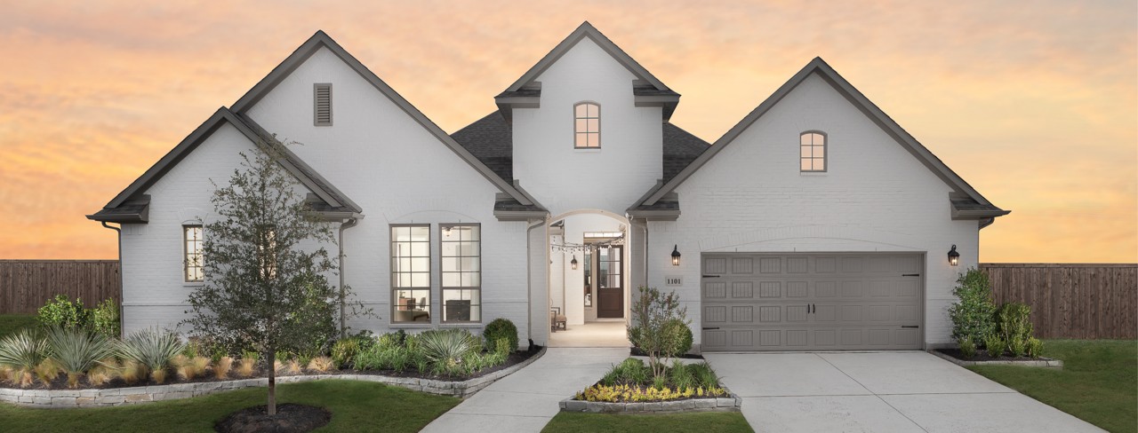 Street view of new home painted white with steep gables, attached garage, and lush landscaping at sunset.