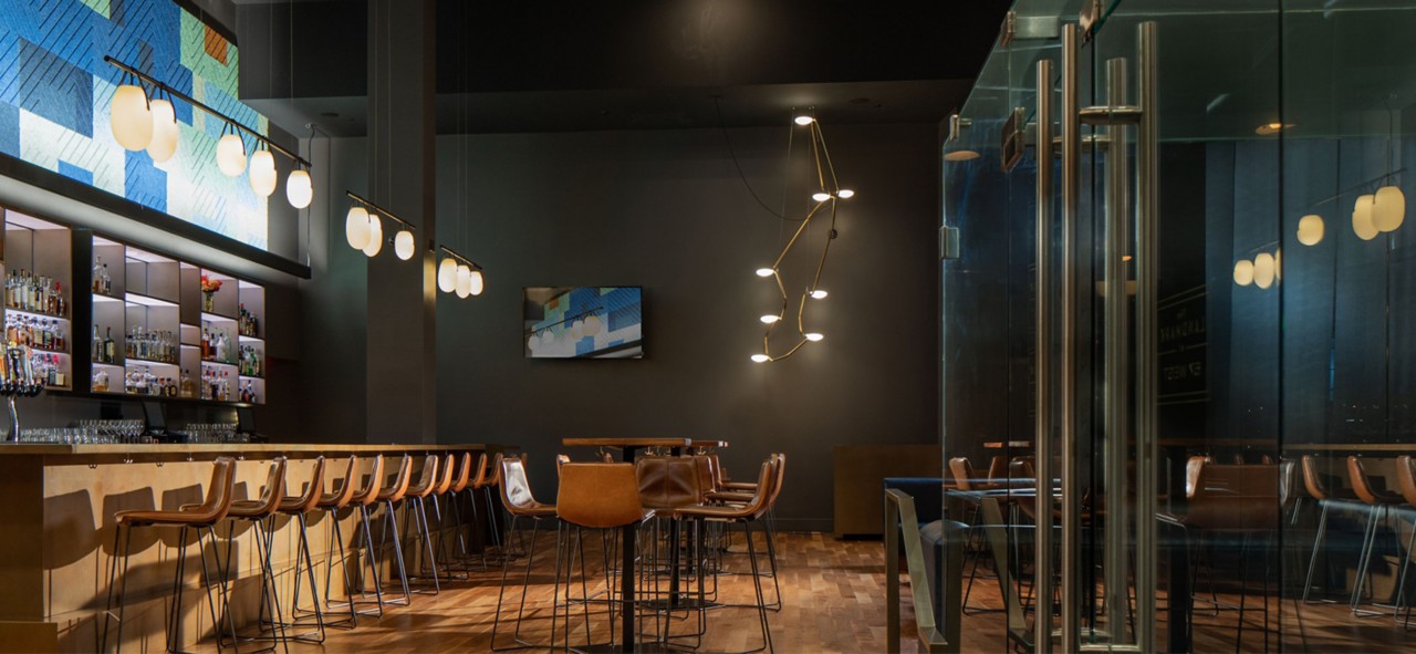 Moody dark interior of restaurant with wood floors, high ceilings, modern lighting, and leather chairs at wood tables and bar.