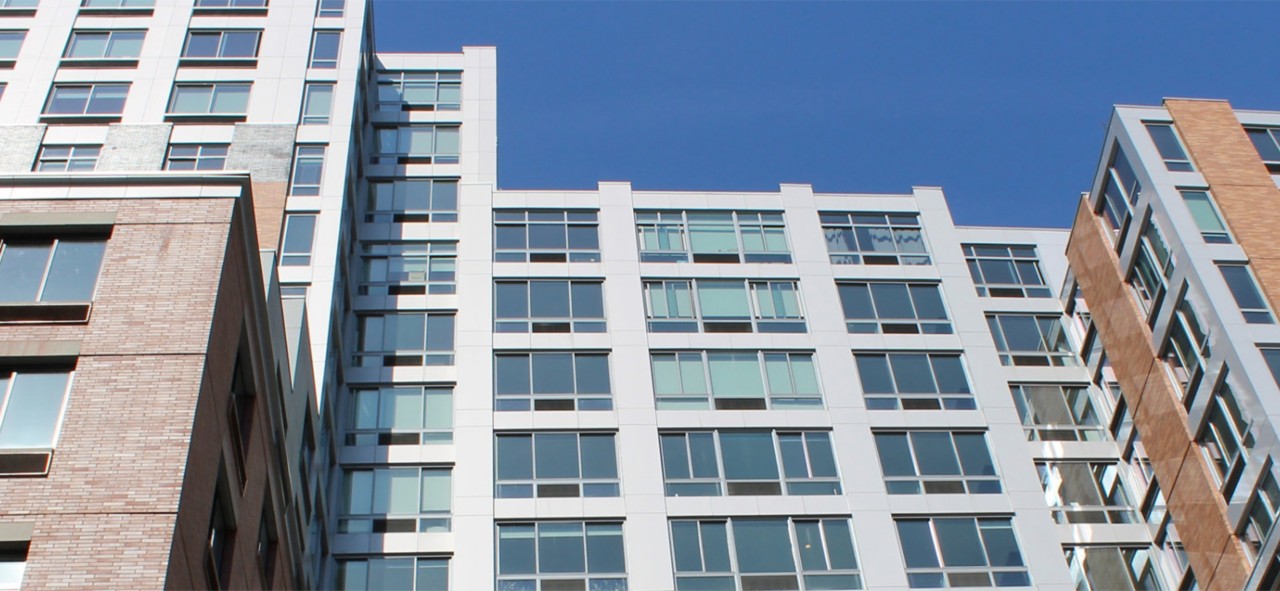 Exterior shot looking up at a city apartment building with brick and metal exterior, wide windows, and clear blue sky above.