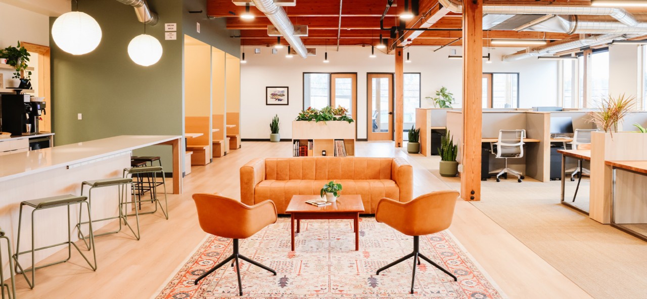 Open concept office space with light wood flooring, exposed wood beams, kitchen counter under globe lights and walled-off booths at left, sofa and chairs in center, and cubicle workspaces to the right.