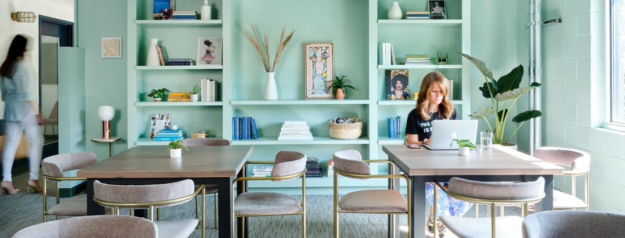 Interior of the Lola coworking space, with light blue-green walls and built-in shelving full of books and decor, with a woman working on a laptop at one of two modern worktables in foreground.