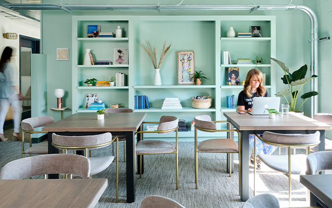 Interior of the Lola coworking space, with light blue-green walls and built-in shelving full of books and decor, with a woman working on a laptop at one of two modern worktables in foreground.