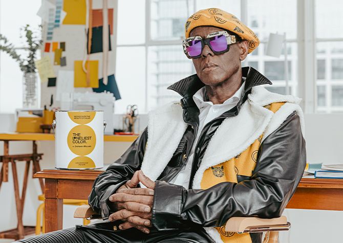 Dapper Dan wearing his custom-designed apparel and mirror sunglasses, seated in front of a wood desk with a can of paint beside him.