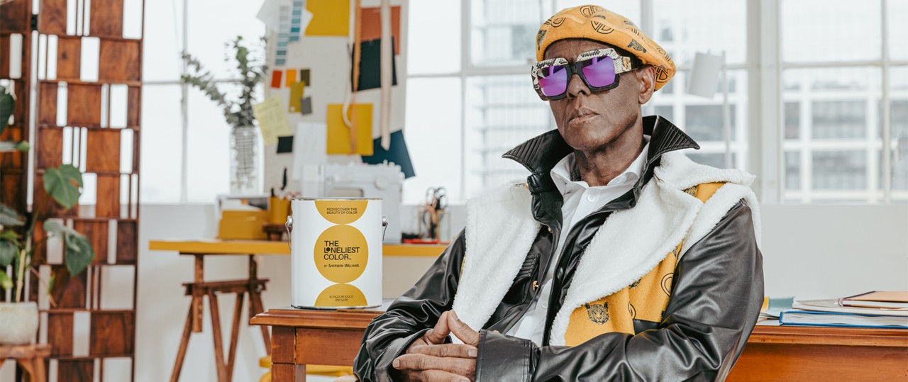 Dapper Dan wearing his custom-designed apparel and mirror sunglasses, seated in front of a wood desk with a can of paint beside him.