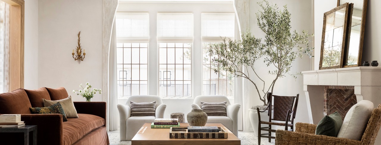 Elegant living room with a brown sofa, white armchairs, large windows, coffee table with books, and a stone fireplace with mirrors and a backdrop of walls painted Greek Villa by Sherwin-Williams.