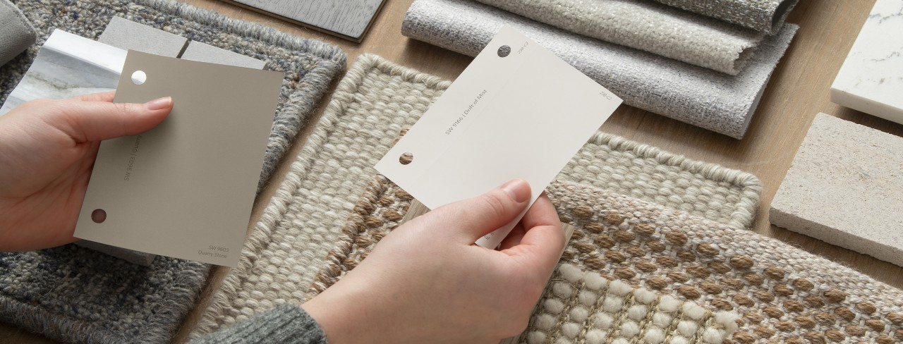 Fabric and textile swatches in a neutral color scheme and hands holding sample color chips in Sherwin-Williams colors Quarry Stone and Drift of Mist.