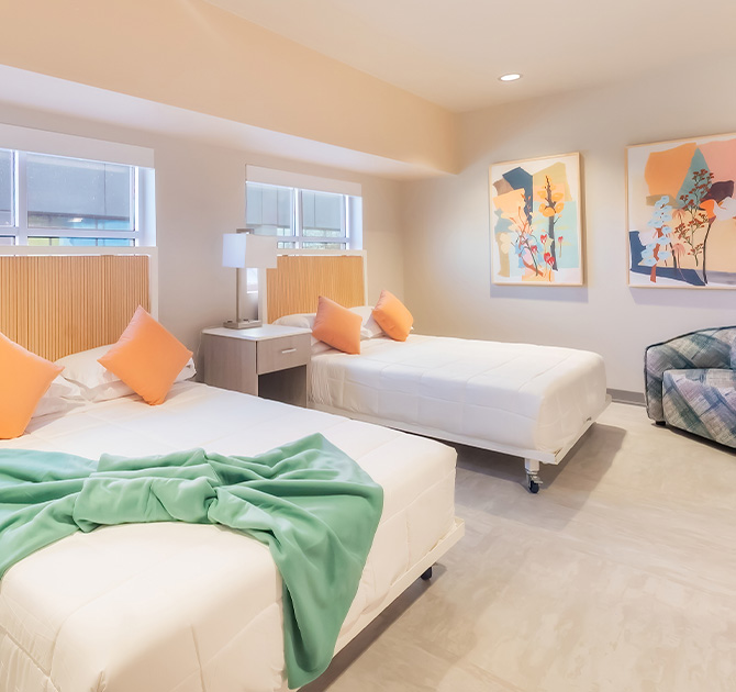 A Ronald McDonald House bedroom with two double beds, white bedding, orange pillows, a central nightstand with a lamp, abstract art on walls painted in Sherwin-Williams Pediment, and a blue plaid armchair.