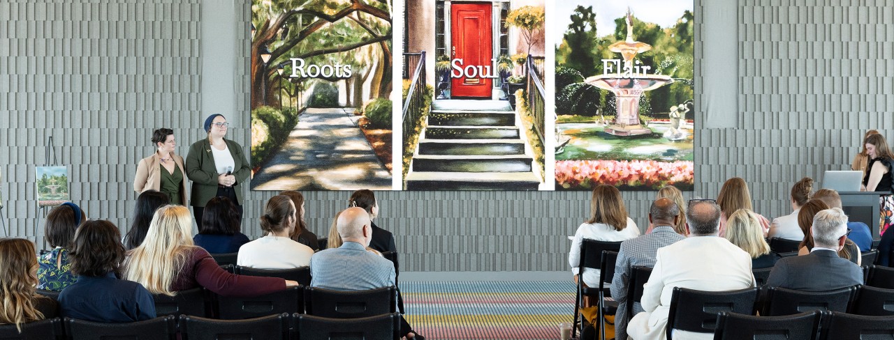 A group of people seated facing a projected image of three artworks displayed on a screen, representing the Roots, Soul, and Flair palettes of the Colors of Savannah project, with presenters standing nearby.