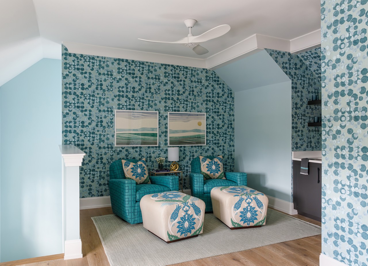 A sitting area on a second-floor landing with patterned blue wallpaper, two teal armchairs with floral footrests, and landscape art prints featuring Watery and Pure White paint colors by Sherwin-Williams.