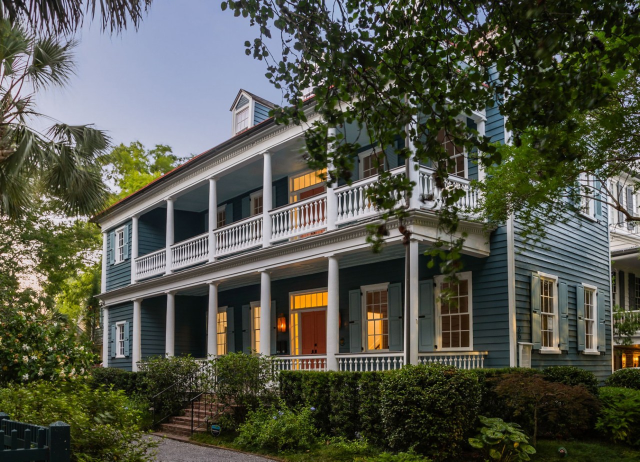 A two-story blue house with white trim, featuring front porches supported by columns, surrounded by greenery.