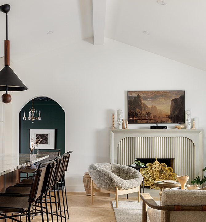 A modern open-concept living space with a kitchen bar, pendant lighting, vaulted ceilings, and a cozy living room with a fireplace and landscape painting.