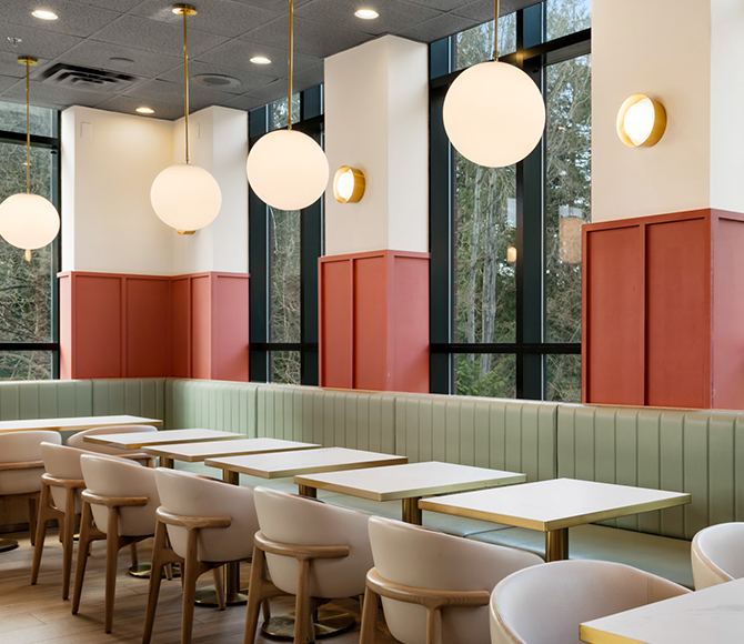Modern restaurant interior with white tables, light green banquettes around perimeter, beige chairs, large windows, and round pendant lights.