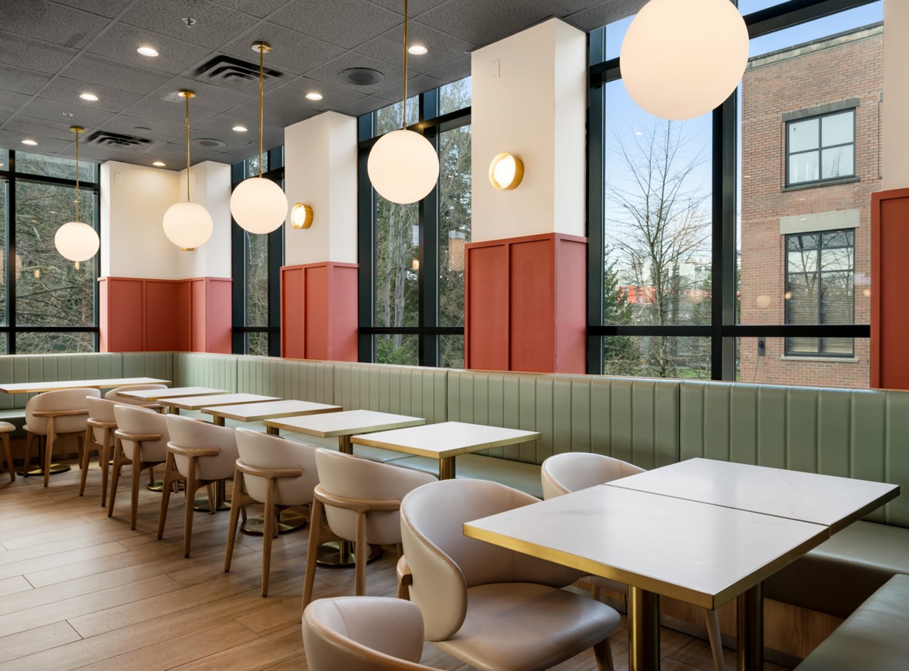 Modern restaurant interior with white tables, light green banquettes around perimeter, beige chairs, large windows, and round pendant lights.