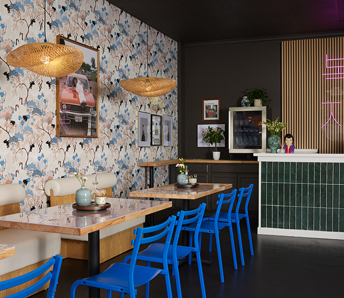 A café interior with blue chairs, wooden tables, floral wallpaper, and a green tiled bar.