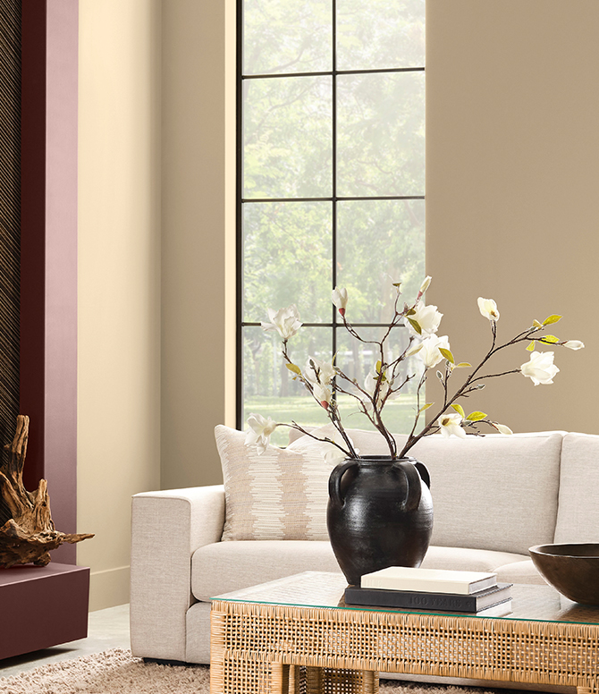 Modern living room with Universal Khaki walls, Dark Auburn painted fireplace, beige sofa, dark vase with flowers, and a glass-topped coffee table.