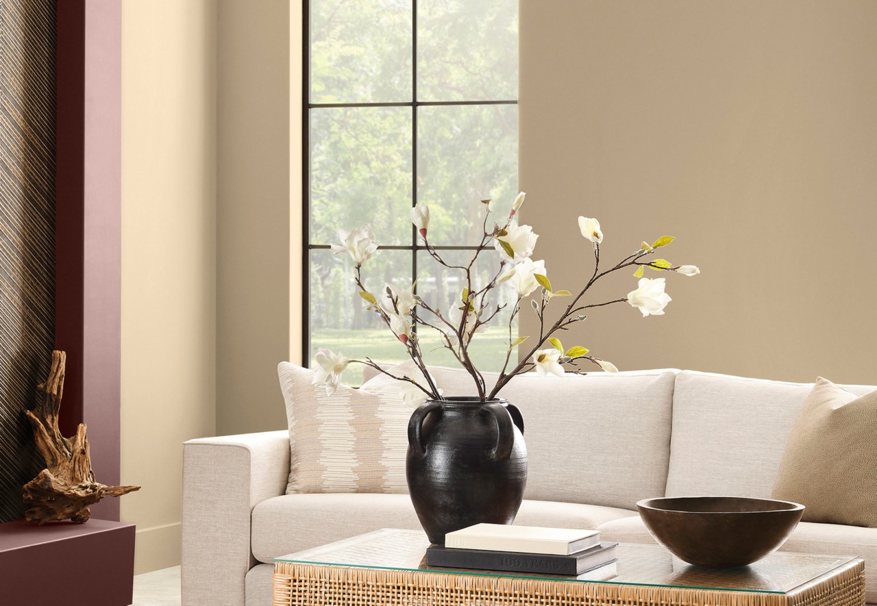 Modern living room with Universal Khaki walls, Dark Auburn painted fireplace, beige sofa, dark vase with flowers, and a glass-topped coffee table.