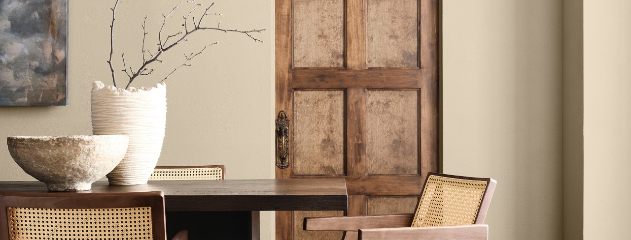 Interior with a dark wooden table, light wooden chairs, ceramic bowl, and vase with branches in front of a Universal Khaki wall and wooden door.