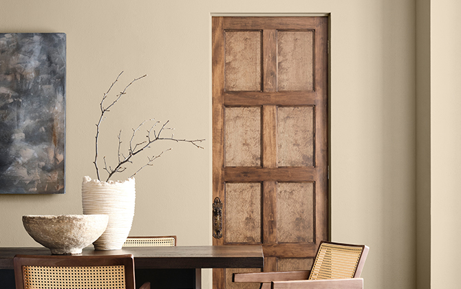 Interior with a dark wooden table, light wooden chairs, ceramic bowl, and vase with branches in front of a Universal Khaki wall and wooden door.