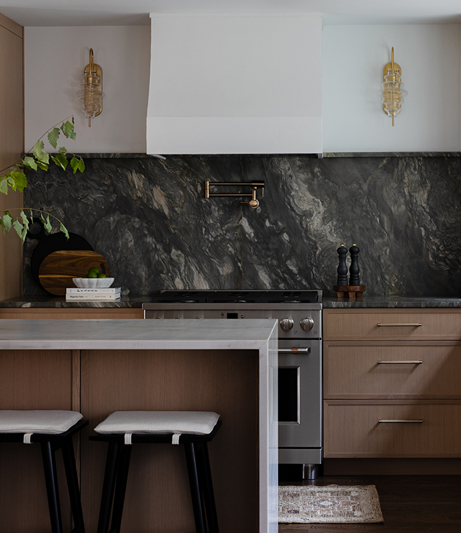 Modern kitchen with light wood cabinetry and a dark stone backsplash, featuring a vase with branches, stainless steel oven, and warm lighting. 