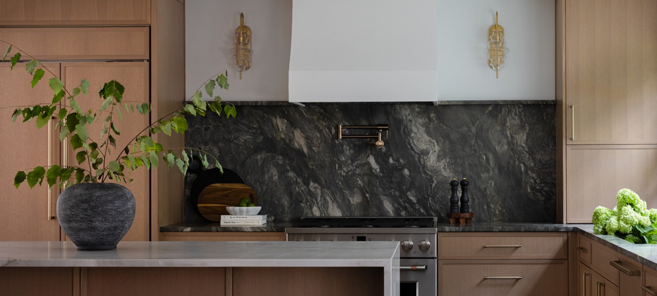 Modern kitchen with light wood cabinetry and a dark stone backsplash, featuring a vase with branches, stainless steel oven, and warm lighting. 