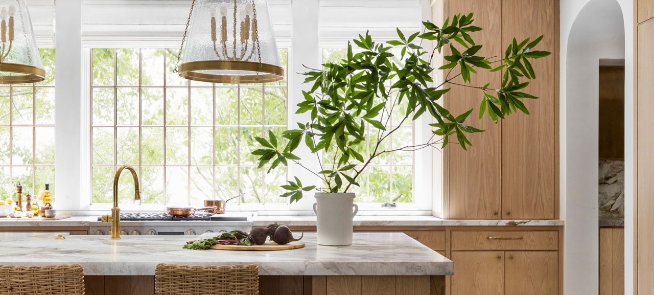 A bright kitchen with marble island, potted plant, wicker stools, wooden cabinets, and large windows. 